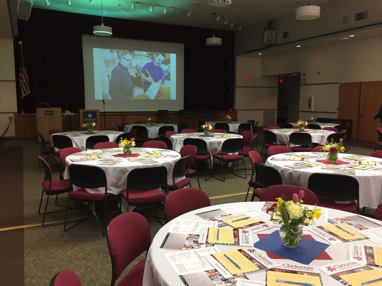 Many tables with red chairs, centerpieces, display material and a slideshow on the projector at the end of the room, in Gregory Forum at the Oregon CIty campus